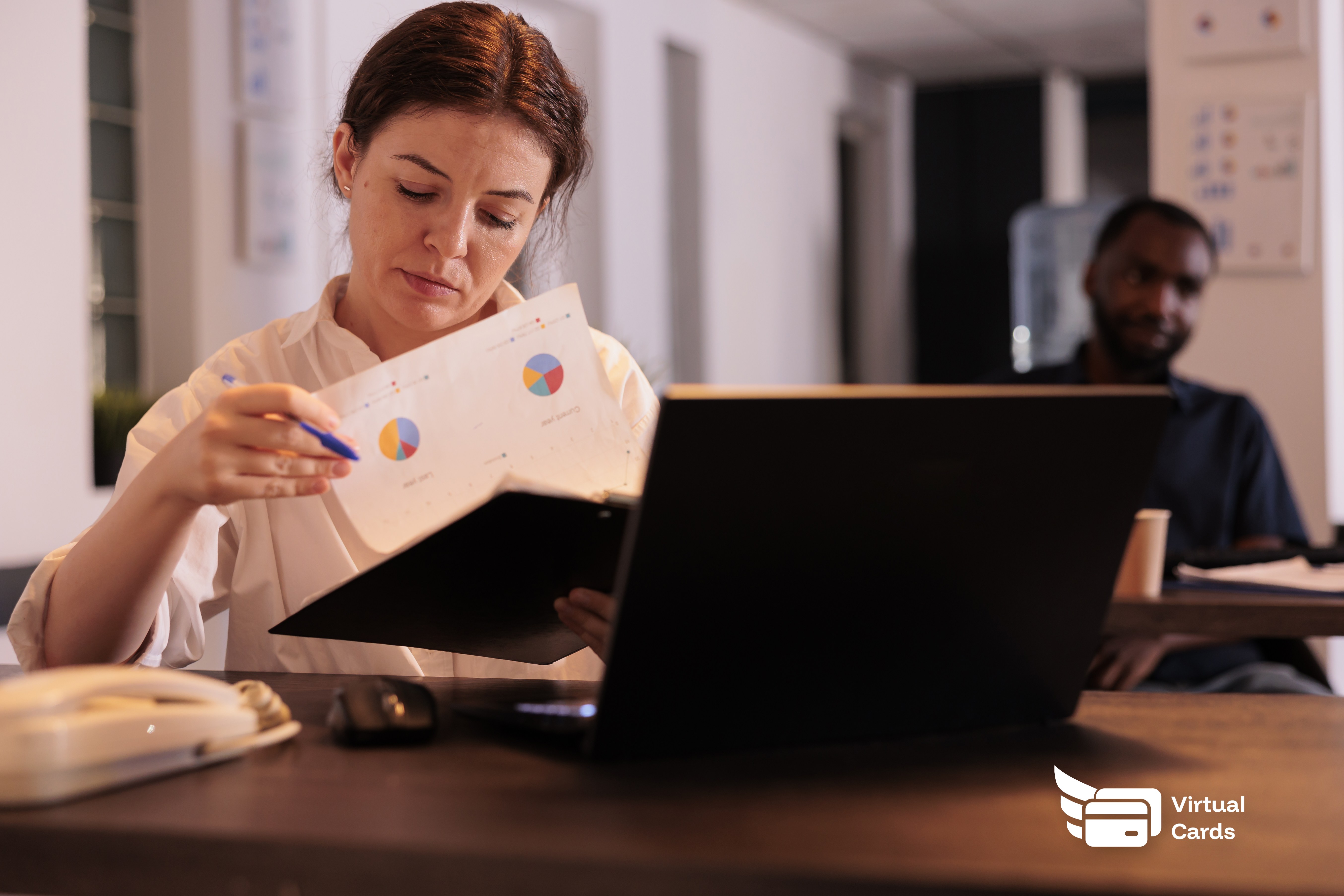 A project manager at a desk reviewing a laptop with project budgets and spending reports, a virtual Visa card in a wallet app on the screen.
