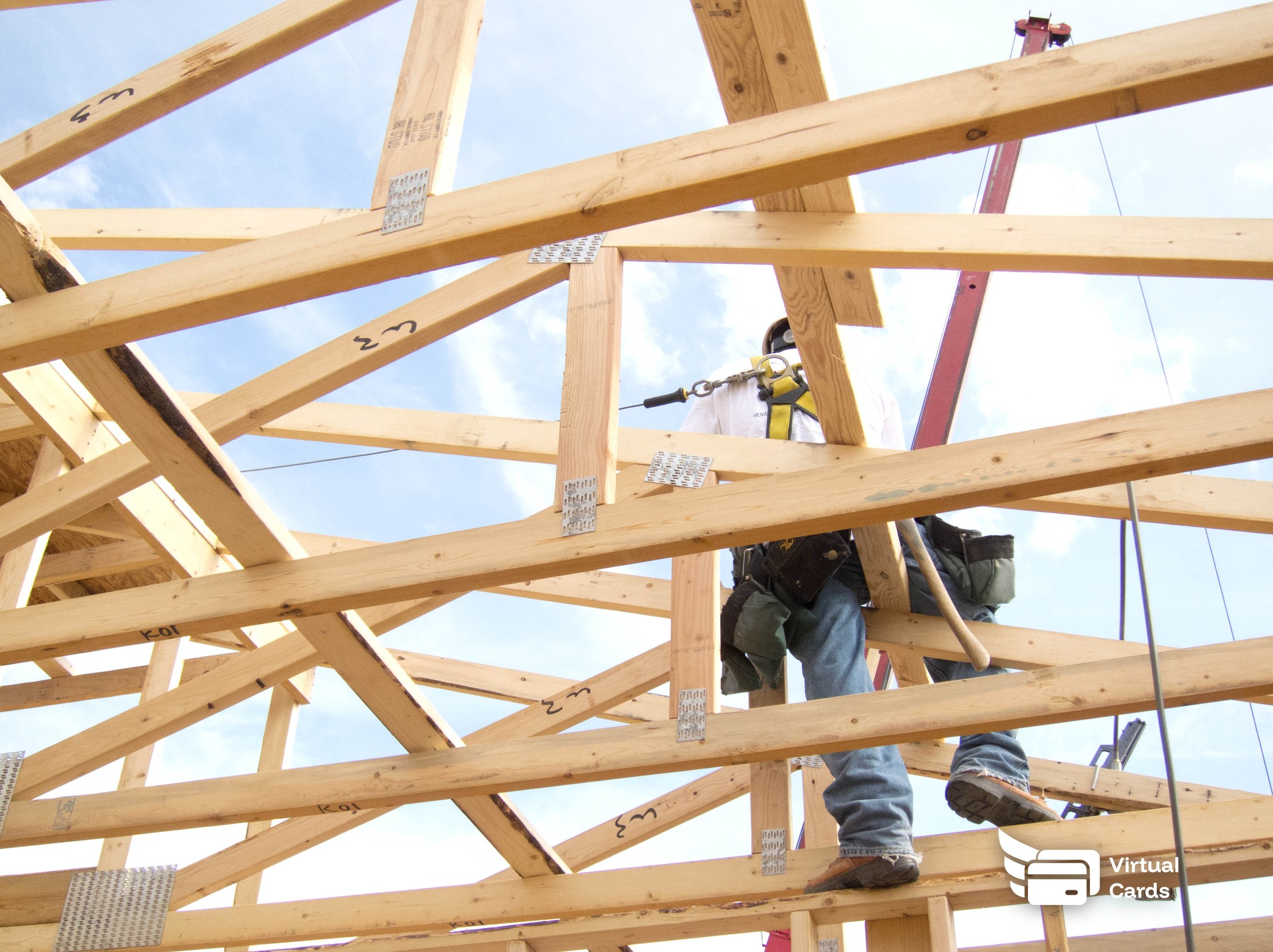 A residential construction worker on a wood roof truss frame wearing a fall-prevention harness, representing a residential construction crew that carries individual virtual cards on the job.